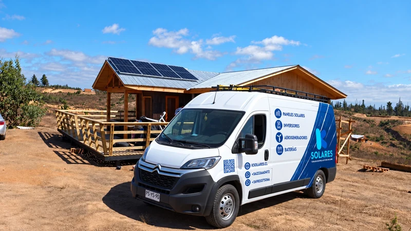 Vehículo de K Solares frente a una vivienda de madera con paneles en la cubierta.