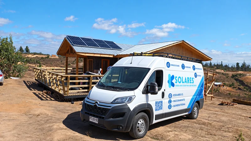Vehículo de K Solares frente a una vivienda de madera con paneles en la cubierta.